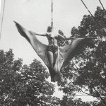female circus performer, dressed as a butterfly, hanging from a wire by her teeth